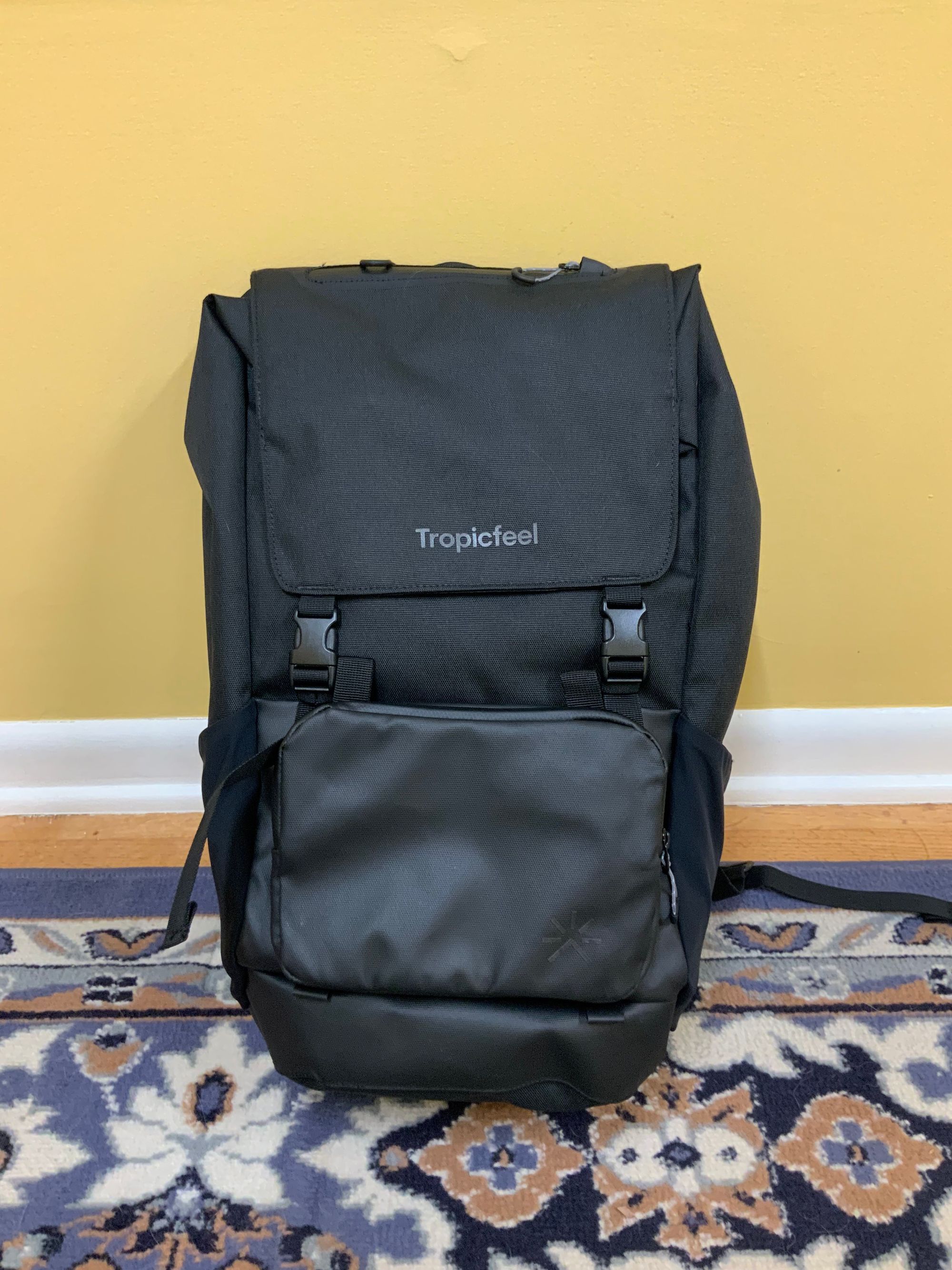 Backpack sitting on the floor. It’s all black with a subtle Tropicfeel logo mark.