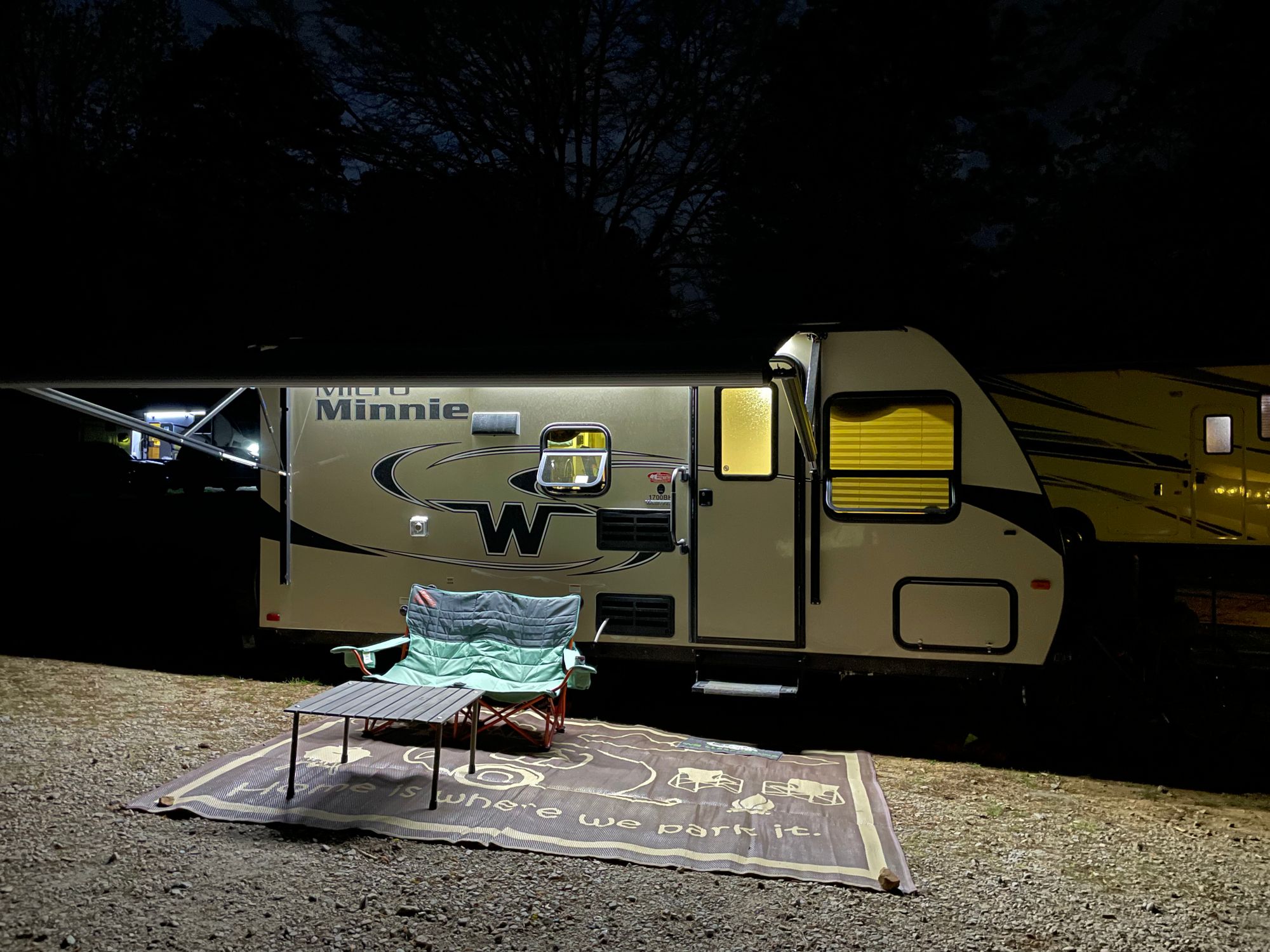 Travel trailer lit up at night. There's a rug outside with a loveseat camp chair and a small table.