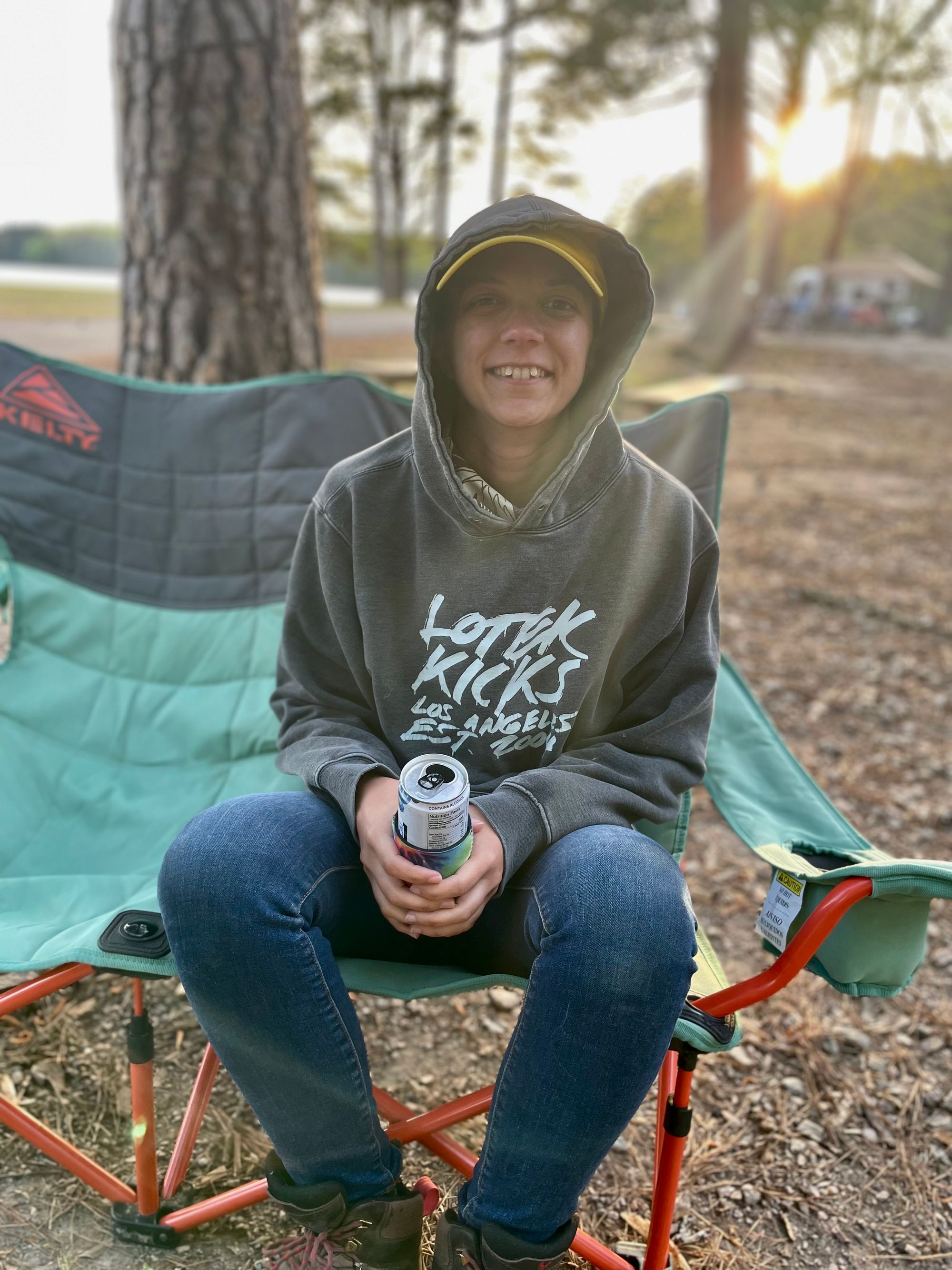 White woman sitting in loveseat camping chair. She's smiling, holding a can, and wearing a hoodie with the hood up.
