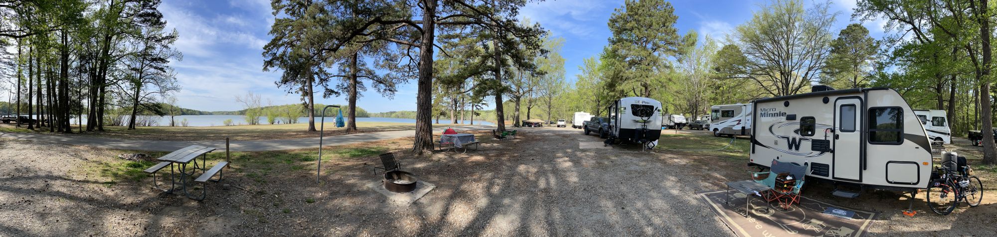 Panoramic view of the lake, trees, picnic tables, fire pits, neighbors' trailer, and the Minniebago.