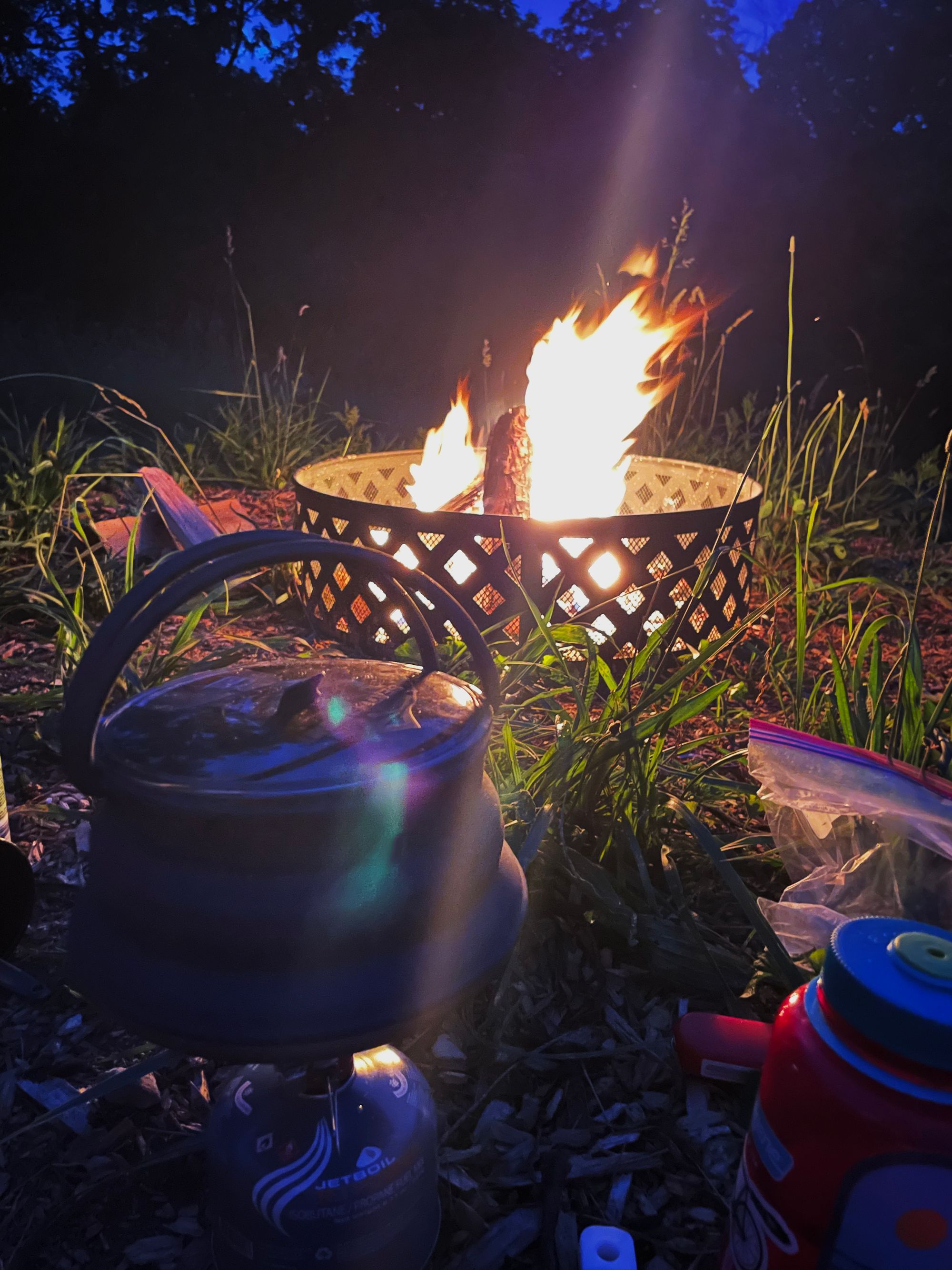 A collapsable kettle sits on a stove on the ground. A fire can be seen in the background.