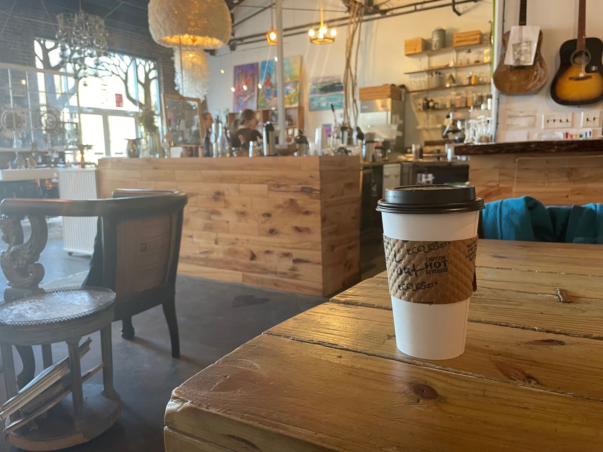 Coffee cup sitting on a wooden table inside a coffee shop. Guitars hang on the wall, and there are lots of trinkets on shelves in the background.