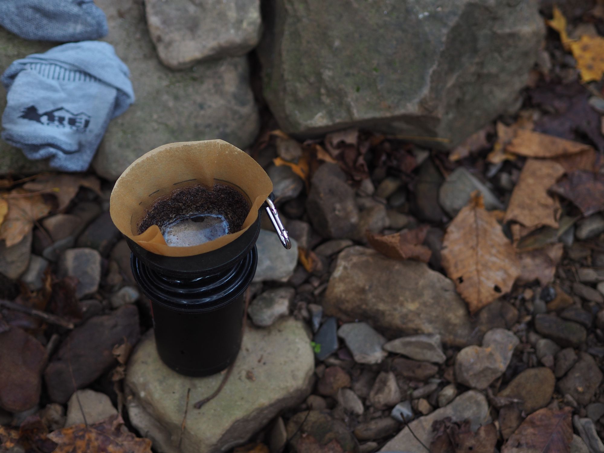 Coffee thermos sitting on rocks with a pour-over filter full of coffee grounds and water on top.