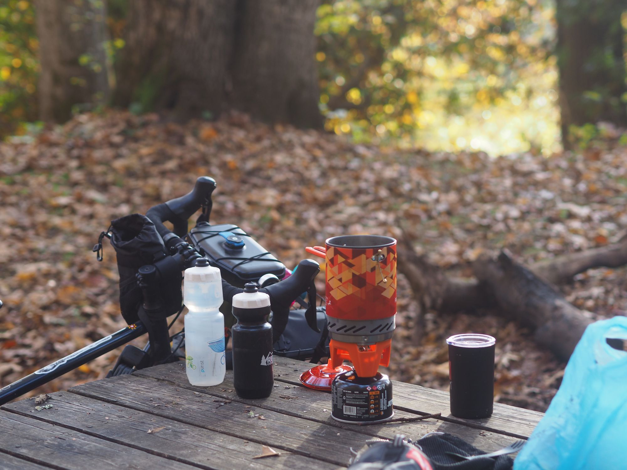 Camping kettle and fuel canister sitting on top of a picnic table in the forest with a bicycle leaning against the table in the background.