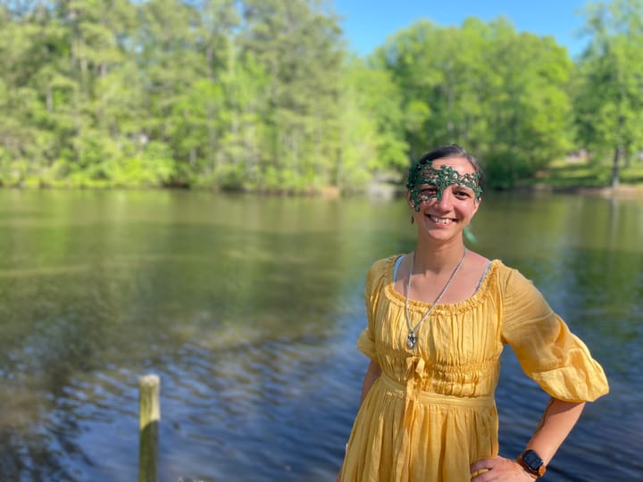 Autumn standing in front of a lake. She's wearing a yellow linen dress, silver necklace, and green half mask.
